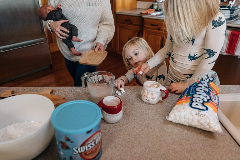 Mom holds newborn while two girls count out marshmallows for their hot chocolate 
