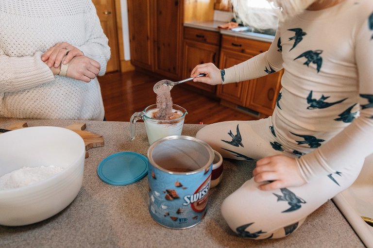 Girl dumps a spoonful of powdered hot chocolate mix in cup of milk. 