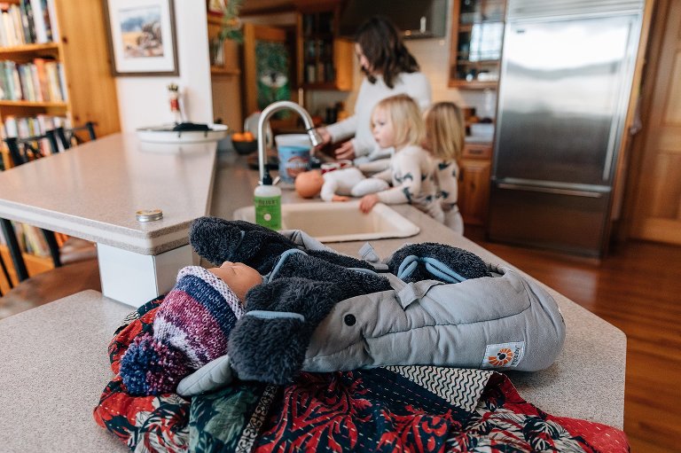 Newborn baby is asleep on the kitchen counter while Mom and sisters prepare hot chocolate in the background. 
