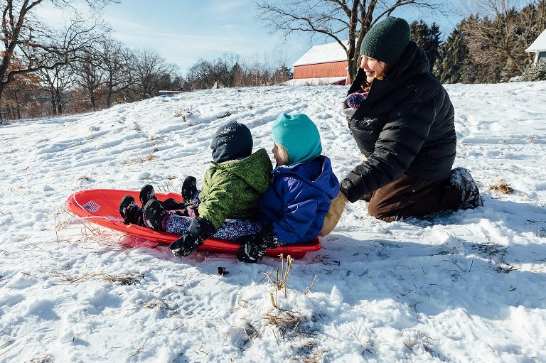 Mom, who is wearing her newborn baby under her winter coat, gerts ready to push her two daughters in a sled. 