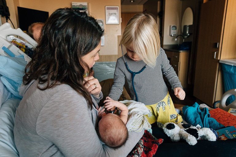 Preschool girl uses stethoscope to listen to newborn sibling's heart at the hospital. Mom holds newborn in the hospital photo. 