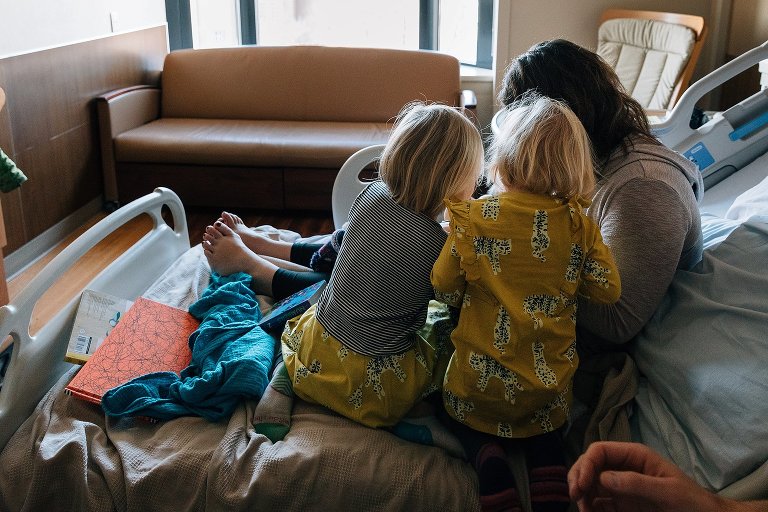 Two girls sit on a hospital with back to camera as the look at newborn sibling with mom. 