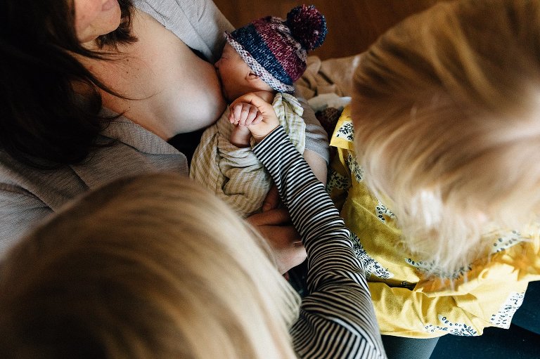 Top down view. Mom nurses and one girl holds newborn's hand while another sister looks on. 