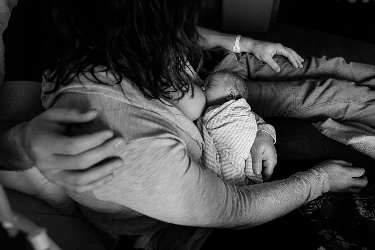 Black and white. Mom nurses newborn while Dad puts arm around Mom. 