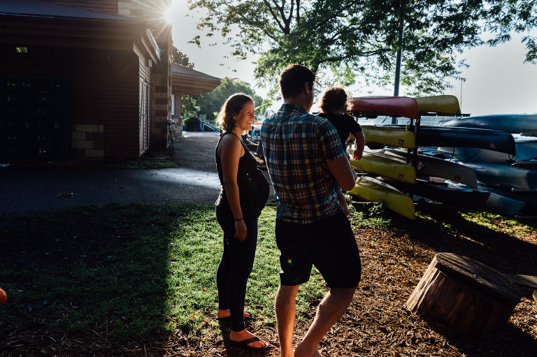 Pregnant woman stands in sunlight at a boat house. 