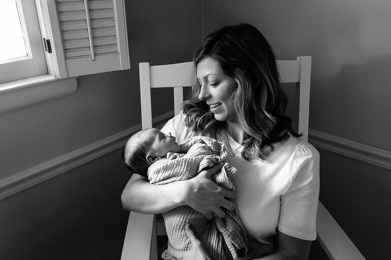 mothers hold her alert newborn in a rocking chair in front of a window