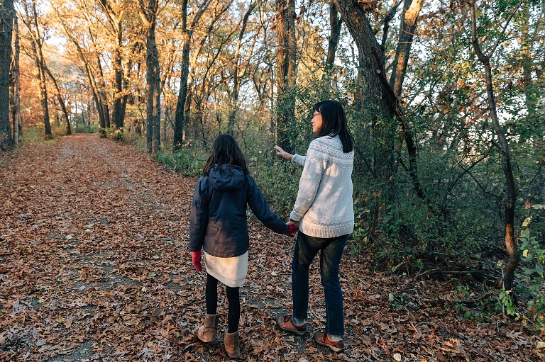 Mothers and daughter walk down an autumn path with light coming through the trees at the end. Mother is gesturing toward the light with her hands.
