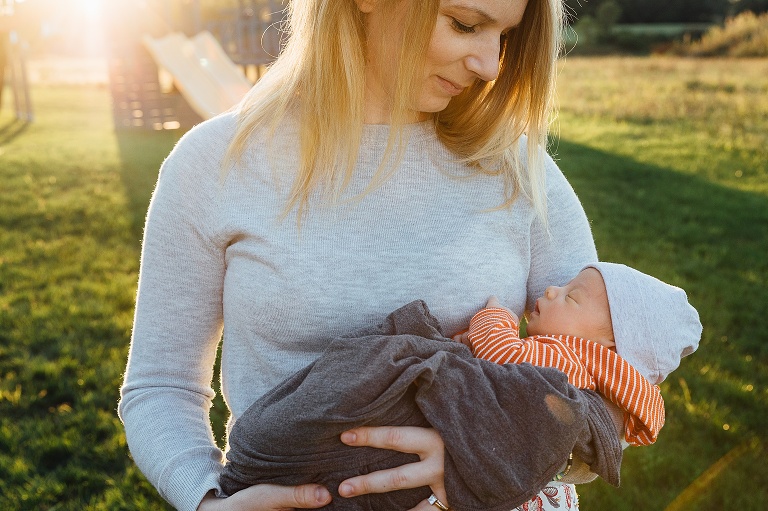 Late evening golden light flares behind young mother looking sweetly at her sleeping newborn in her arms.