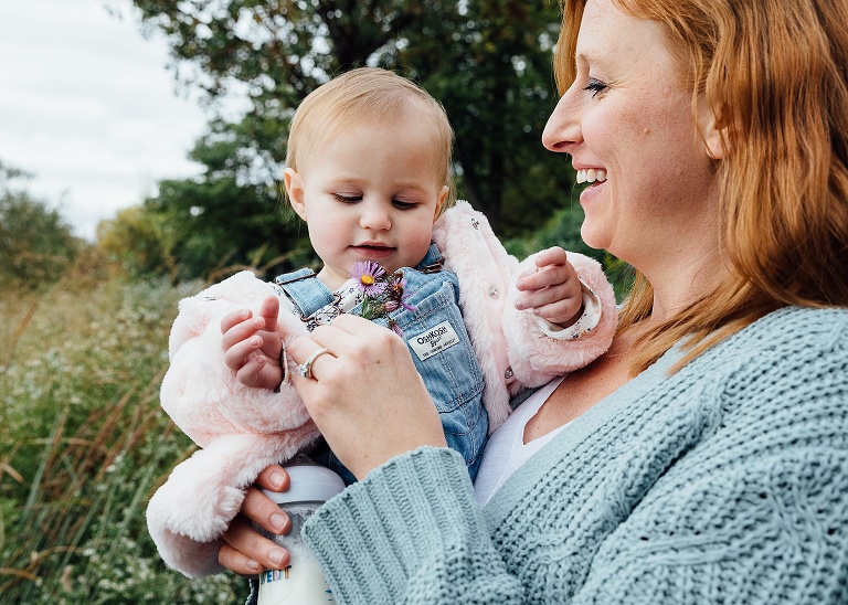 Mother holds toddler daughter in arms while handing her a purple aster flower. Daughter looks curious and smiling.