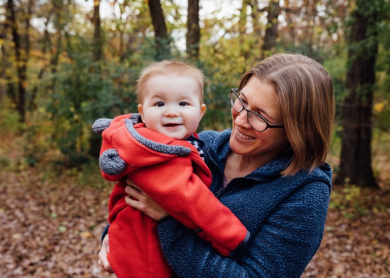 Mother smiles at infant son who is smiling at the camera on a fall day. 