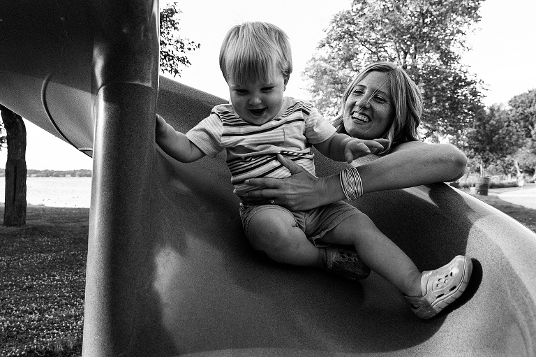 Mothers laughs as toddler son slides down curly slide. Black and white. 