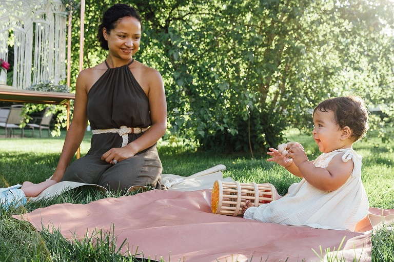 Mothers smile at toddler girl who laughing on a blanket outside. Green trees and green grass in background.