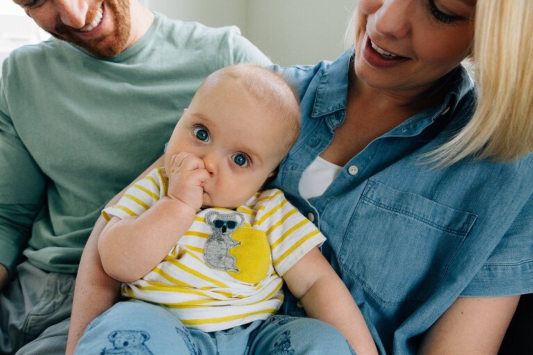 Baby sucks thumb while sitting on mom and dad's laps during Family Photography in MIddleton