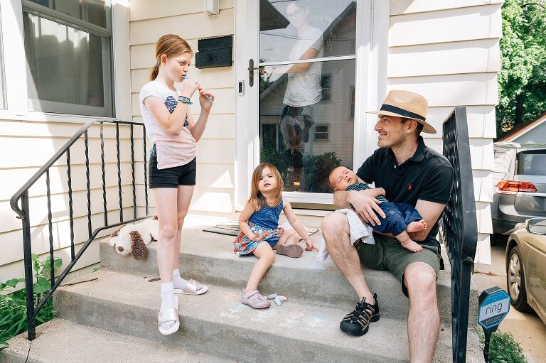 Front porch scene. Dad holding baby. Sister eating Freezer Pop. Toddler drawing with chalk. Mom exiting the house through the front door.
