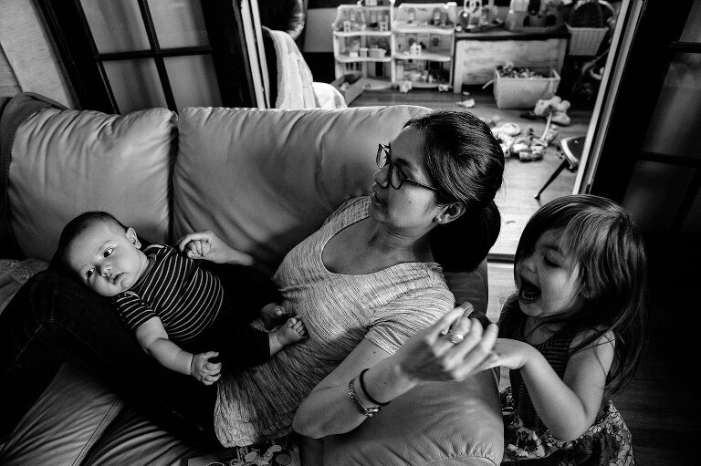 Black and white. Mom lies on the couch holding infant son on her lap and passing something to her toddler daughter behind her. 