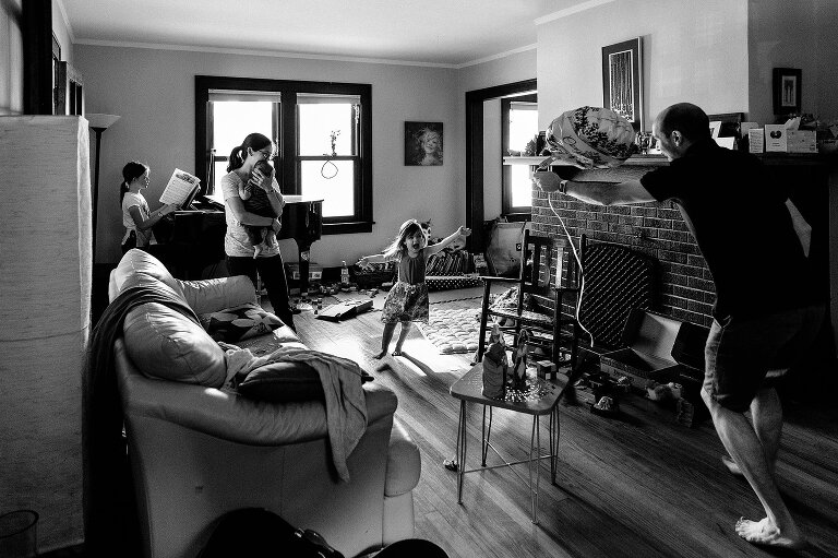 Black and white. Living room scene--girl and dad running toward each other, older girl playing the piano, mom holds infant. 