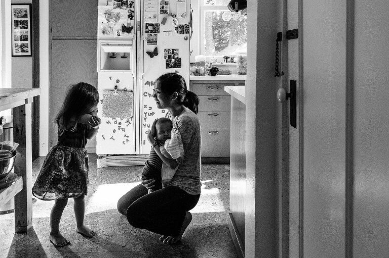 black and white. Mom and preschool girl talk in the kitchen while mom holds infant an ordinary summer Saturday morning