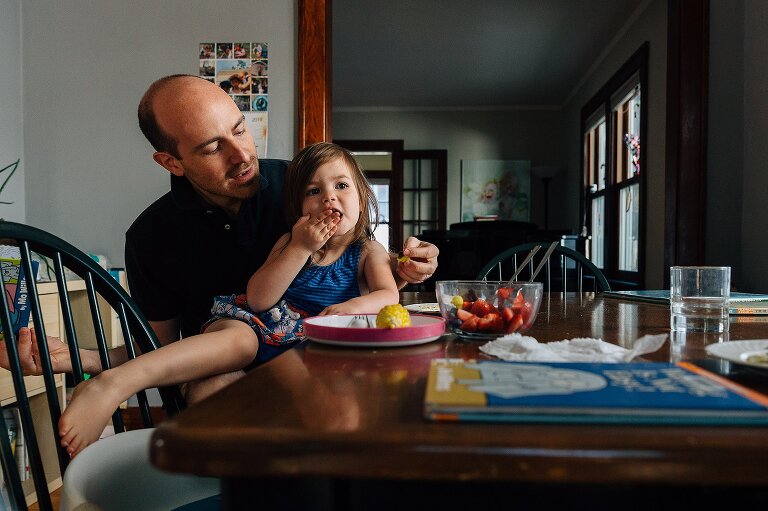 young girl sits on her dad's lap eating breakfast on an ordinary summer saturday mornning