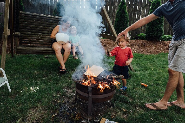 Dad holds young son's head as son approach smoky fire to add kindling. Mom and older son sit on swing beyond them eating popcorn. 