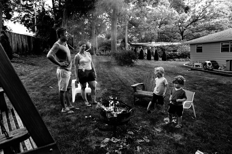 Black and white. Family of 4 make funny faces at each other in backyard campfire during the summer.