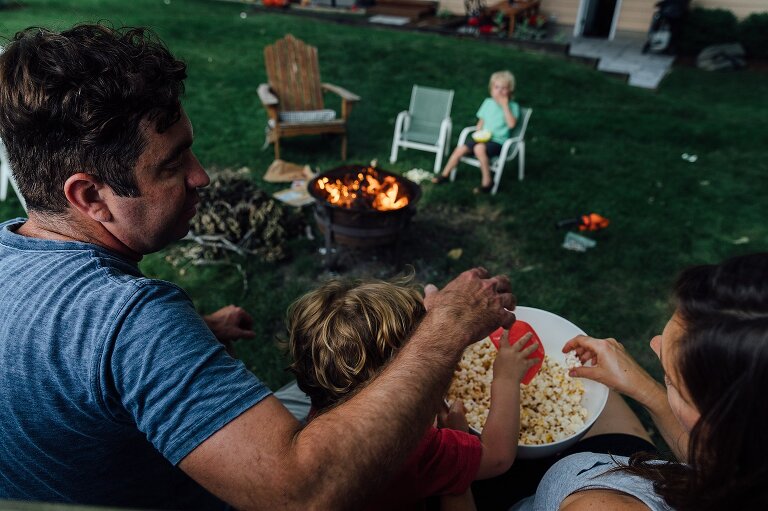 Family all reaches for popcorn at the same time in front of a backyard campfire. 