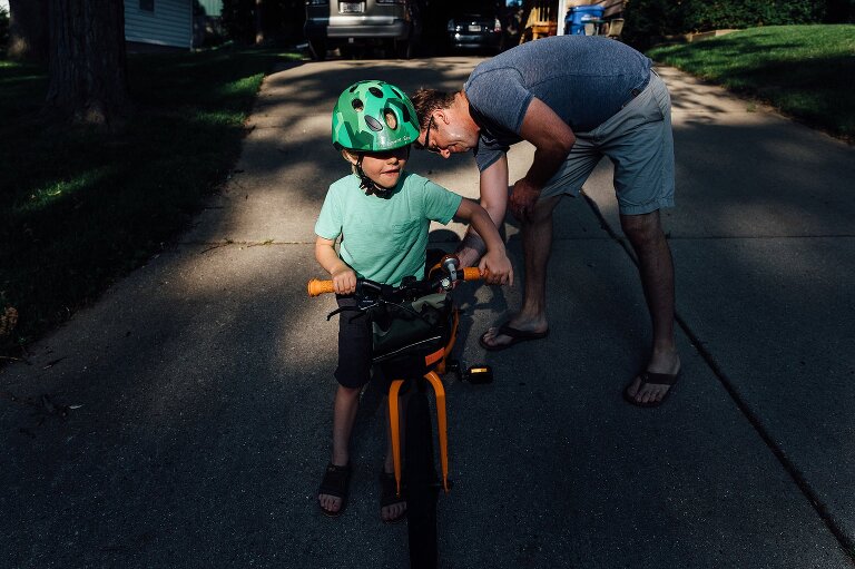 Boy stradels bike in a pocket of light while dad checks bike tires. 