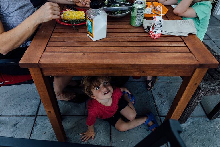 Boy sits under the family table during dinnertime. He looks up through the slats. 