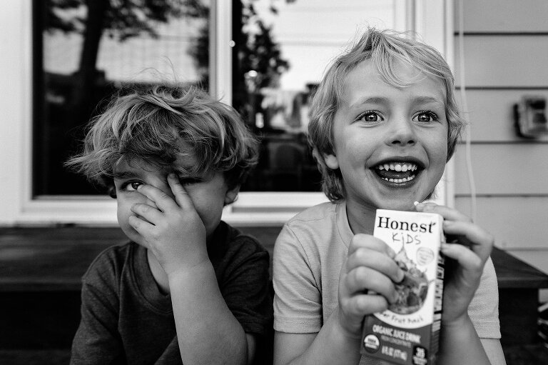black and white. Two young boys sit outside, close up. One boy drinks from juice box, one boy covering his face with his fingers. 