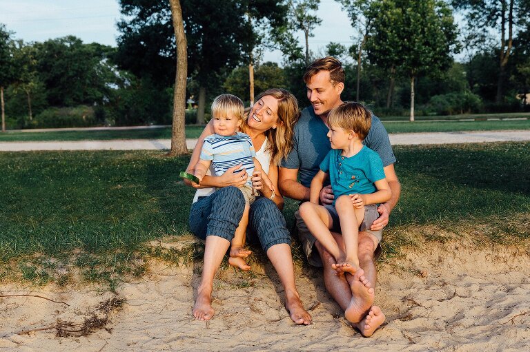 Family of four sits in the sand at the beach. They are engaged with each other and smiling.