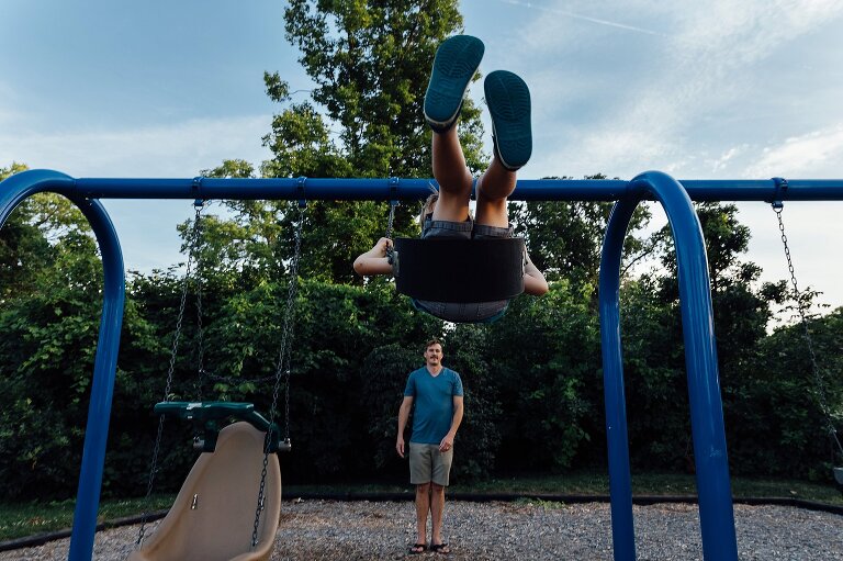 Dad smiles as son swings on a swing. Focus on son's blue shoes that are high in the sky. 