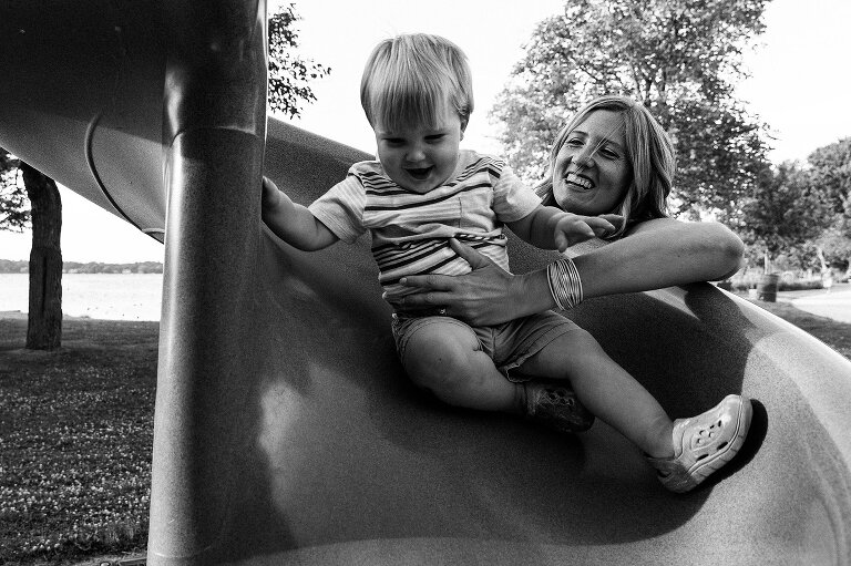 Black and white. Mom smiles as she catches her toddler son coming down a curvy slide.