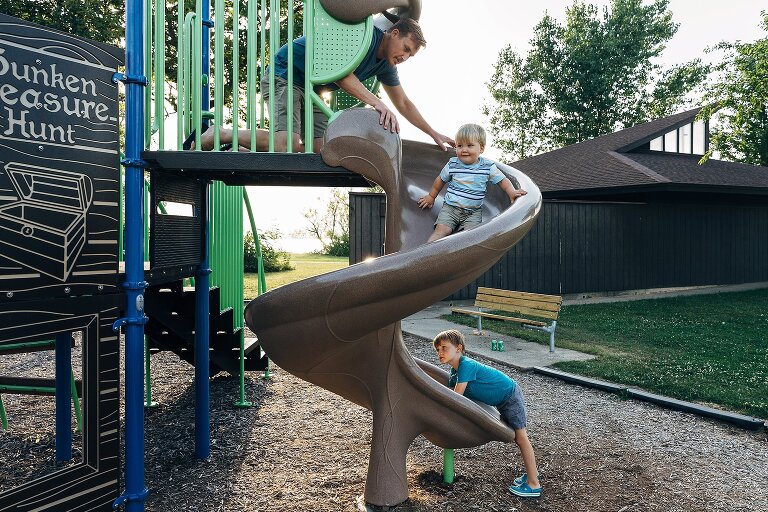 Dad helps toddler go down a slide at the top. Older preschooler waits for brother's safety at the bottom. 