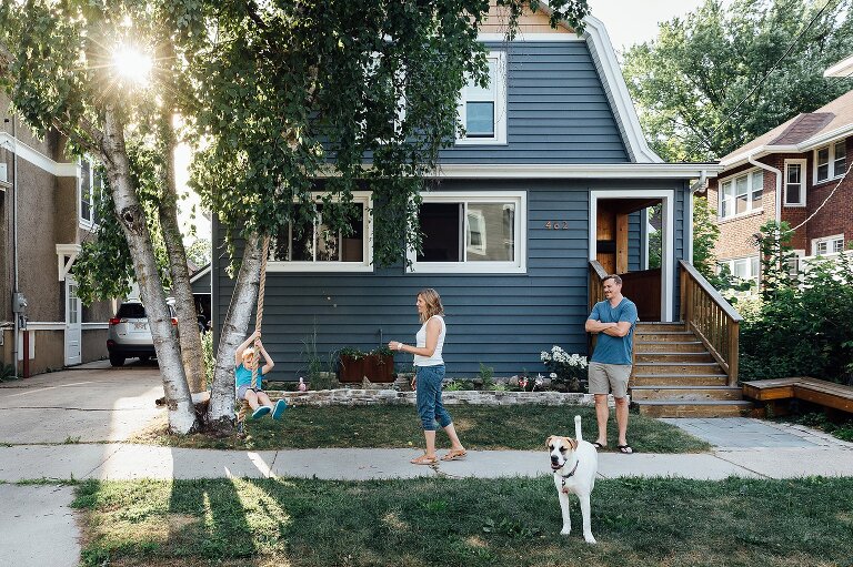 Family of 3 and dog in front of a blue house on a sunny day. Boy swings from tree rope.