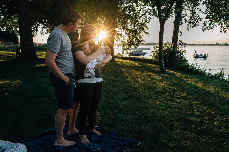 Mom holds swaddled newborn in the backyard on the lake with sun shining through the trees 