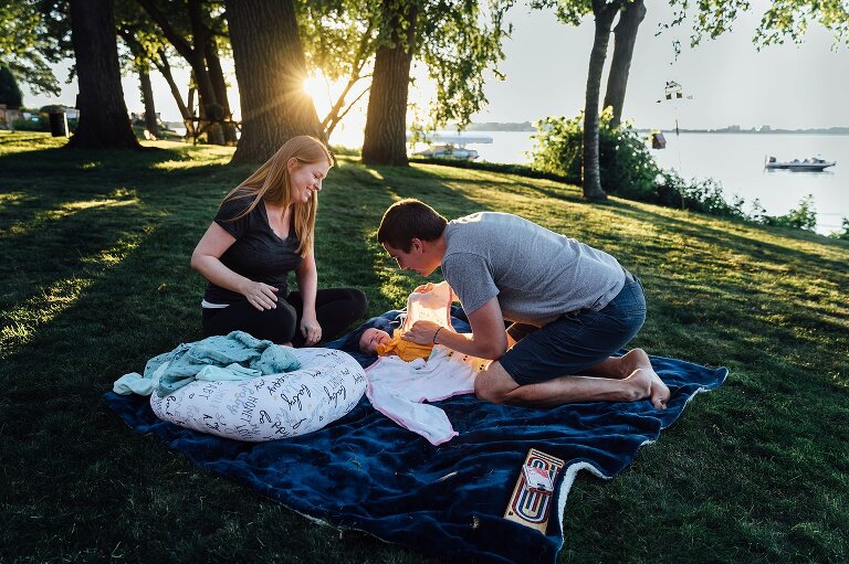 Dad swaddles newborn while mom watches while in the backyard on the lake 