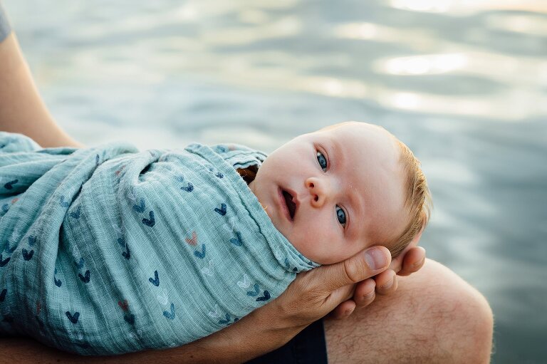 Newborn makes eye contact with camera while swaddled in blanket and lake in the background