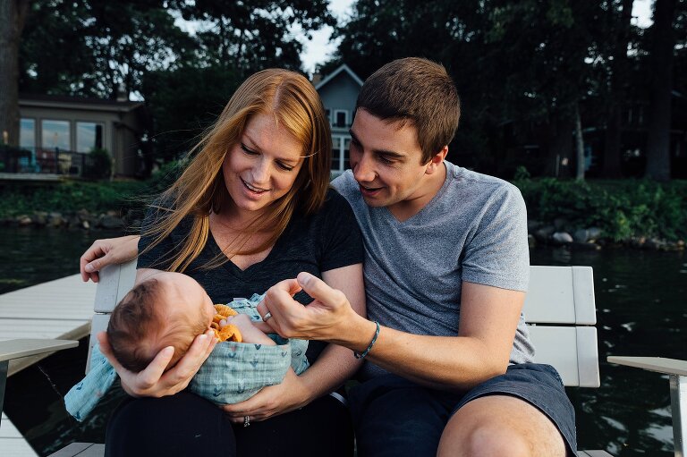 Mom holds newborn while Dad holds newborn's finger in the backyard on the lake