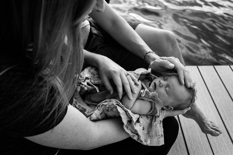 B/W. Focus on sleeping newborn with two parents touching her with lake in the background. 