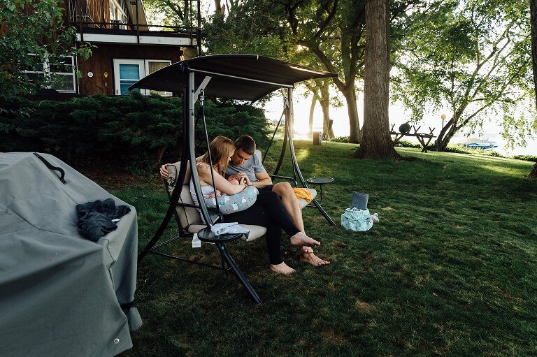 Two parents look at their nursing newborn while sitting on a swing in their backyard on the lake