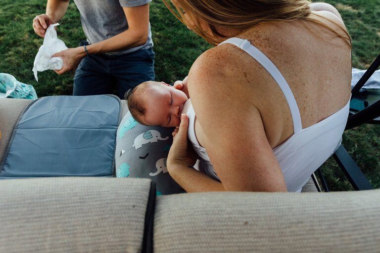 Mom nurses newborn on a backyard swing while dad rolls up diaper in the background