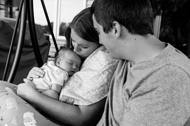 blw. Mom kisses sleeping newborn's head while Dad looks on