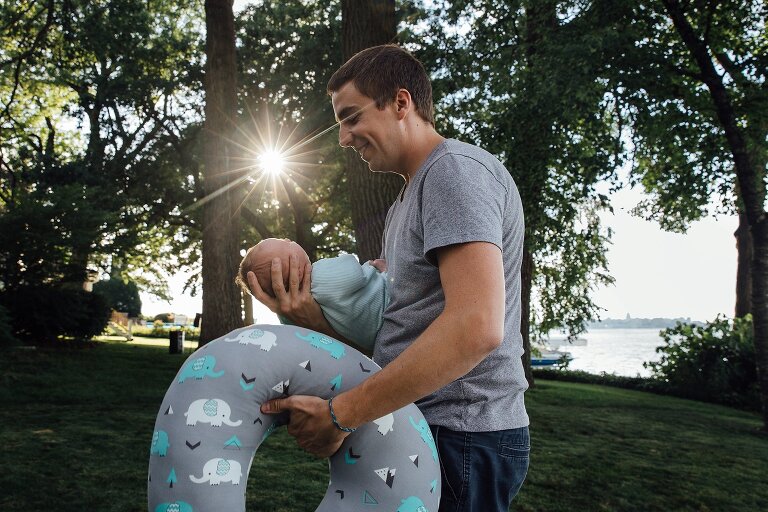 Dad holds newborn and carries Boppy as he walks through his backyard on the lake with sun shining