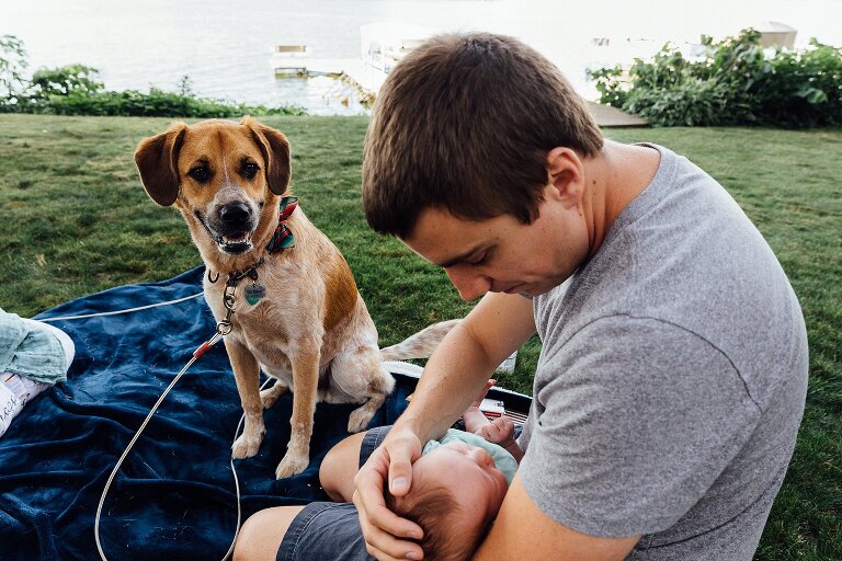 Dad cuddles newborn in their backyard on the lake while dog looks on