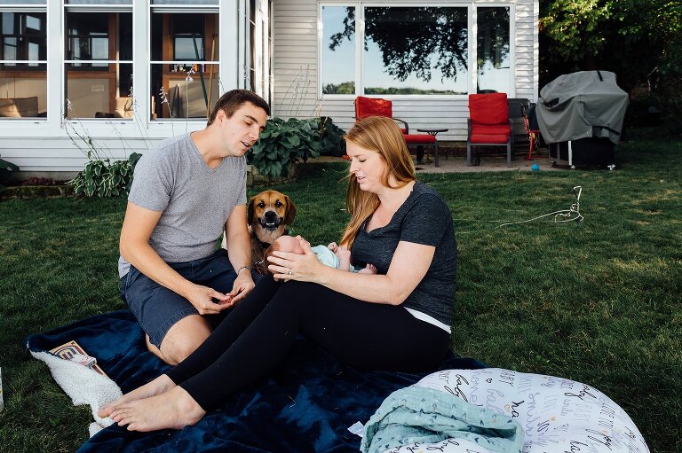 Two parents, newborn, and a smiling dog sit in their backyard overlooking the lake.