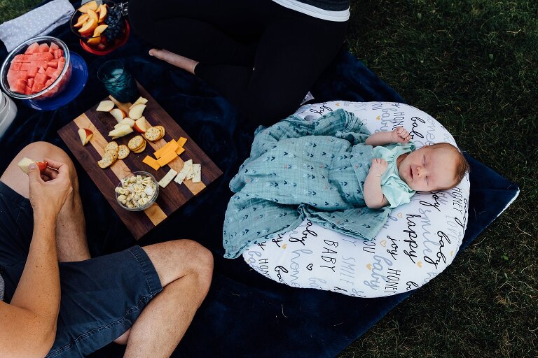 Newborn sleeps on a Boppy in the grass while parents eat a picnic spread next to her. 