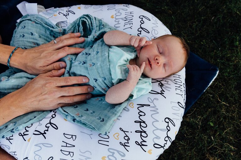 Dad's hands tuck in a blanket around newborn. Outside in the grass on a boppy.