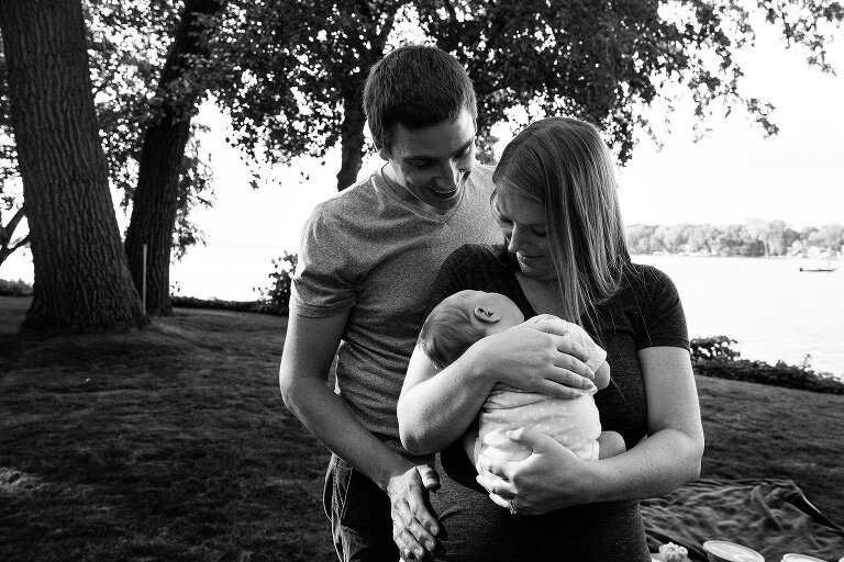 black and white. Dad stands behind mom and newborn and smiles at them.