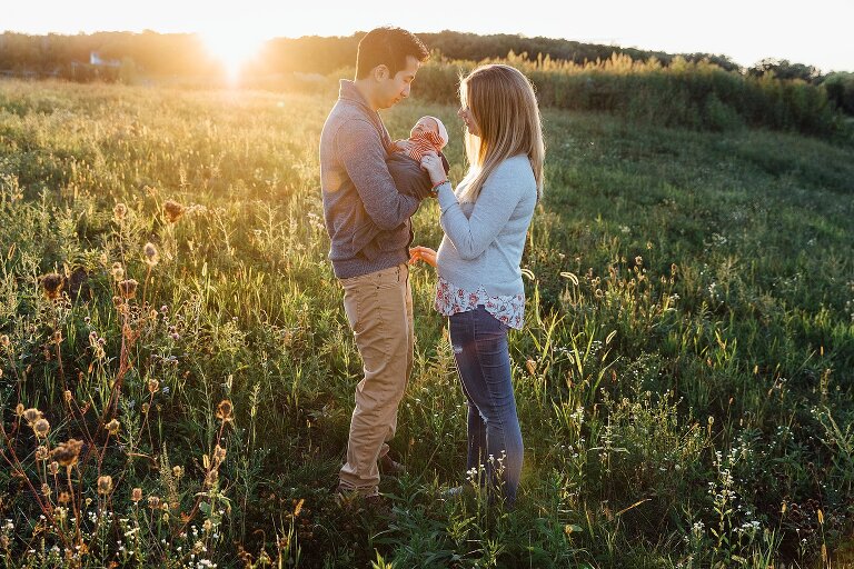 Dad holds newborn while mom touches his cheek while standing in a field in their backyard.