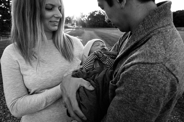 Black and white. Newborn is between two parents on a walk with sunshine in the distance. 