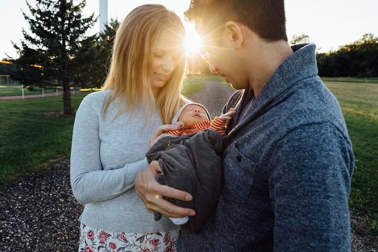 Mom and Dad hold newborn on a walk. Sun shines between them.